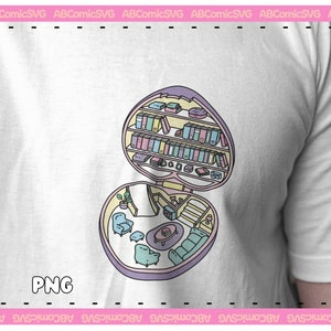 Puede incluir: Camiseta blanca con un juguete en forma de coraz&oacute;n abierto, que revela una habitaci&oacute;n en miniatura con muebles y una estanter&iacute;a. El juguete es de colores pastel, con el texto "PNG" en el centro.