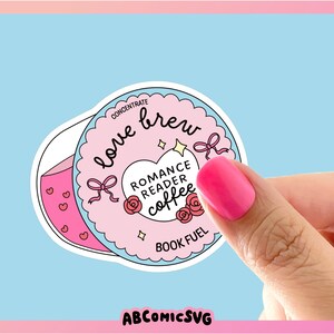 Puede incluir: Una pegatina con un dise&ntilde;o rosa y blanco, con las palabras "love brew" y "Romance Reader Coffee". La pegatina tiene un fondo azul claro e incluye lazos, corazones y rosas. Tambi&eacute;n est&aacute; presente el texto "Book Fuel". La imagen est&aacute; sostenida por una mano con esmalte de u&ntilde;as rosa.
