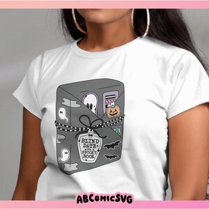 May include: White t-shirt with a graphic of a gray book tied with a black ribbon and decorated with ghosts, bats, and a pumpkin. The text on the book reads "Blind Date with a Spooky Book".