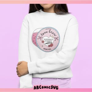 Puede incluir: Sudadera blanca con un gr&aacute;fico. El gr&aacute;fico incluye una taza de caf&eacute; rosa y un dise&ntilde;o circular con el texto "love brew", "Romance Reader Coffee" y "Book Fuel". El dise&ntilde;o incluye corazones, rosas y lazos.