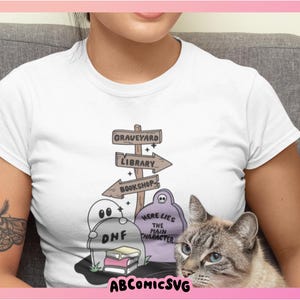 May include: White t-shirt featuring a whimsical graveyard scene with directional signs pointing to a graveyard, library, and bookshop. The design includes cartoon ghosts, books, and the text "Here Lies The Main Character". A cat is in the image.