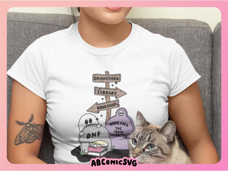 May include: White t-shirt featuring a whimsical graveyard scene with directional signs pointing to a graveyard, library, and bookshop. The design includes cartoon ghosts, books, and the text "Here Lies The Main Character". A cat is in the image.