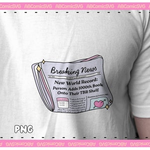 Puede incluir: Una camiseta blanca con una ilustraci&oacute;n rosa y morada de un titular de peri&oacute;dico que dice "Breaking News: New World Record: Person Adds 1000th Book Onto Their TBR Shelf". La ilustraci&oacute;n incluye un coraz&oacute;n rosa con una flecha y brillos.