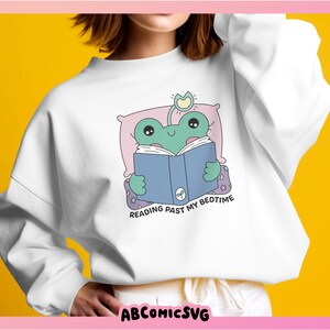 Op de afbeelding: Wit sweatshirt met een groene kikker die in bed een blauw boek leest. De tekst op het sweatshirt luidt "Reading past my bedtime."