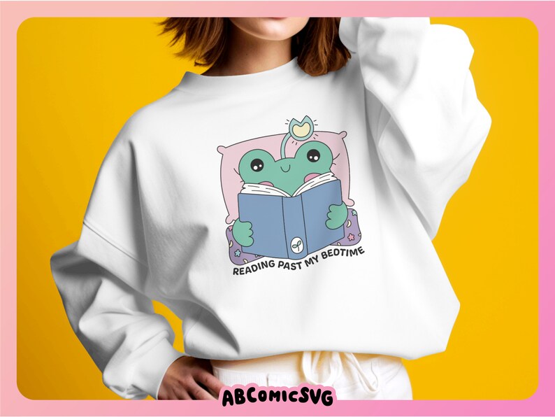 Op de afbeelding: Wit sweatshirt met een groene kikker die in bed een blauw boek leest. De tekst op het sweatshirt luidt "Reading past my bedtime."