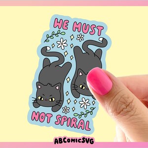 May include: A sticker featuring two gray cartoon cats, one upright and one upside down, on a light blue background. The sticker has the text "WE MUST NOT SPIRAL" in pink. White flowers and green vines decorate the background. The sticker is held by a hand with pink nail polish.