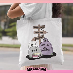 May include: White tote bag featuring a whimsical graveyard scene with tombstones, ghosts, and directional signs pointing to a graveyard, library, and bookshop. The text "Here Lies The Main Character" is on a tombstone. The bag also has the text "ABCOMICSVG".