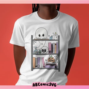 May include: White t-shirt featuring a graphic of a ghost reading a book on a bookshelf decorated with Halloween items. The bookshelf includes a skull, a pumpkin, potions, and candles.