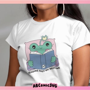Op de afbeelding: Wit T-shirt met een groene kikker die in bed een boek leest. De kikker draagt een blauw boek en heeft een roze kussen. De tekst op het shirt luidt "Reading past my bedtime".