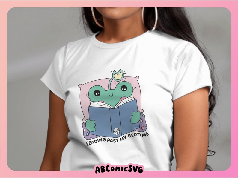 Op de afbeelding: Wit T-shirt met een groene kikker die in bed een boek leest. De kikker draagt een blauw boek en heeft een roze kussen. De tekst op het shirt luidt "Reading past my bedtime".