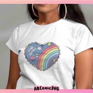 May include: White t-shirt with a rainbow heart design and the text "safe space" in blue lettering. The heart design includes stars, planets, and a rainbow.