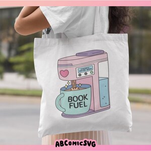 May include: White tote bag featuring a pink coffee maker illustration with a heart and the text "BOOK FUEL" on a blue mug. The coffee maker has the text "ESSENTIAL ENTIRE BOOK". The bag has the text "ABCOMICSVG" at the bottom.