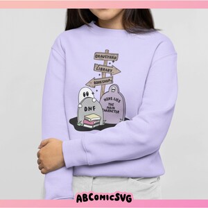 May include: A light purple sweatshirt featuring a graveyard scene with tombstones, a ghost, and directional signs pointing to a graveyard, library, and bookshop. The text "Here Lies The Main Character" is on a tombstone. The design is whimsical and cartoonish.