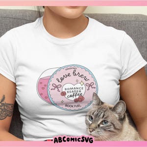 Puede incluir: Camiseta blanca con un gr&aacute;fico circular con las palabras "love brew" y "romance reader coffee". El dise&ntilde;o incluye corazones rosas, lazos y rosas. Un gato gris con ojos azules est&aacute; en primer plano. Tambi&eacute;n se ve el texto "BOOK FUEL".