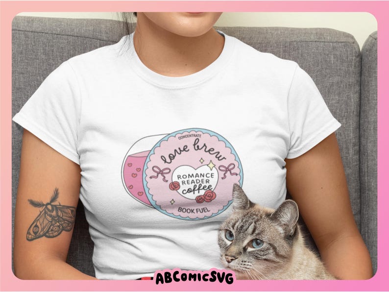 Puede incluir: Camiseta blanca con un gr&aacute;fico circular con las palabras "love brew" y "romance reader coffee". El dise&ntilde;o incluye corazones rosas, lazos y rosas. Un gato gris con ojos azules est&aacute; en primer plano. Tambi&eacute;n se ve el texto "BOOK FUEL".
