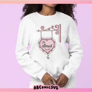 May include: White sweatshirt with a pink heart graphic that says "The Smut Reader" on it. The heart is hanging from a decorative sign.