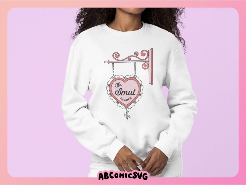 May include: White sweatshirt with a pink heart graphic that says "The Smut Reader" on it. The heart is hanging from a decorative sign.