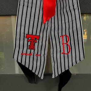 May include: A graduation stole featuring a black and white vertical striped pattern with a bright red ribbon at the top. The left side displays a red embroidered Texas Tech logo with the text wreck 'em, and the right side features a red letter B.