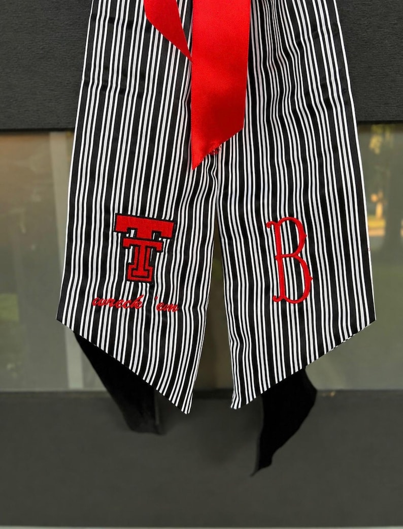 May include: A graduation stole featuring a black and white vertical striped pattern with a bright red ribbon at the top. The left side displays a red embroidered Texas Tech logo with the text wreck 'em, and the right side features a red letter B.