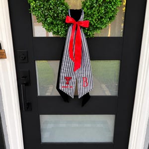May include: A green boxwood wreath adorned with a black and white striped fabric hanger featuring a bright red bow. The fabric displays the red embroidered letters T and B, with the text "wreck 'em" positioned below the letter T in red thread.