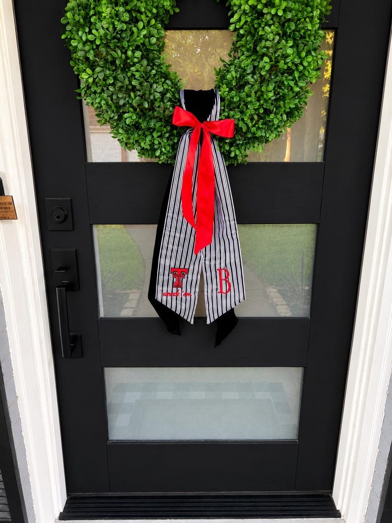 May include: A green boxwood wreath adorned with a black and white striped fabric hanger featuring a bright red bow. The fabric displays the red embroidered letters T and B, with the text "wreck 'em" positioned below the letter T in red thread.