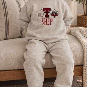 May include: A light heather gray long-sleeve sweatshirt and matching sweatpants set. The sweatshirt features a graphic design with a red letter T, a pointing finger icon, and a cowboy hat icon, with the name SHEP printed in red below the graphics.