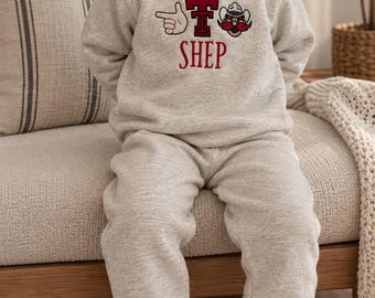Könnte beinhalten: Ein hellgrau meliertes Set aus einem langärmeligen Sweatshirt und einer passenden Jogginghose. Das Sweatshirt zeigt ein grafisches Design mit einem roten Buchstaben T, einem Fingerzeig-Symbol und einem Cowboyhut, darunter der Name SHEP in Rot.