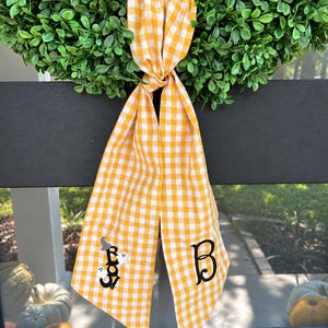 May include: A yellow and white gingham fabric ribbon tied in a knot, featuring an embroidered black letter B on one tail and the word Boo with two small white ghosts and a grey witch hat on the other tail, hanging from a green wreath.