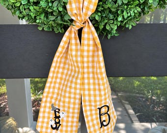 Könnte beinhalten: Ein gelb-weiß kariertes Stoffband mit Knoten, das auf einem Ende den gestickten schwarzen Buchstaben B und auf dem anderen Ende das Wort Boo mit zwei kleinen weißen Geistern und einem grauen Hexenhut zeigt, hängend an einem grünen Kranz.