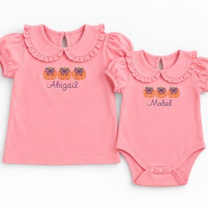 May include: A pink short-sleeved shirt and a matching pink bodysuit, both featuring a ruffled Peter Pan collar and a keyhole back. Each garment is embroidered with three orange pumpkins topped with purple bows and the names Abigail and Mabel.