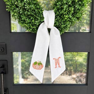 May include: A green boxwood wreath adorned with a white fabric ribbon tied in a knot. The left ribbon tail features an embroidered orange pumpkin with a green stem, while the right ribbon tail displays an embroidered orange letter W monogram.