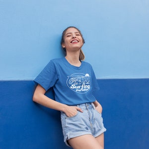 K&ouml;nnte beinhalten: Eine Frau tr&auml;gt ein blaues T-Shirt mit einem wei&szlig;en Aufdruck, auf dem "Surfing Paradise" steht, und blaue Jeans-Shorts. Sie steht vor einer blauen Wand.