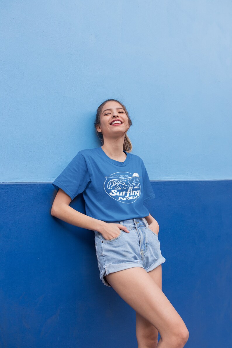 K&ouml;nnte beinhalten: Eine Frau tr&auml;gt ein blaues T-Shirt mit einem wei&szlig;en Aufdruck, auf dem "Surfing Paradise" steht, und blaue Jeans-Shorts. Sie steht vor einer blauen Wand.