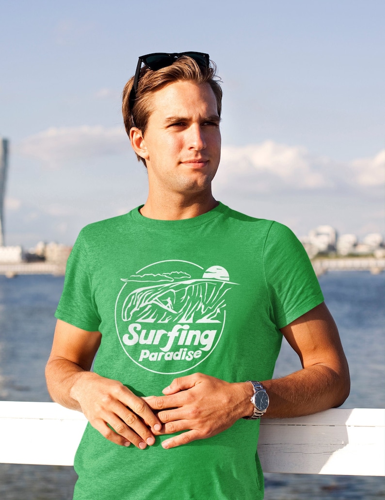 K&ouml;nnte beinhalten: Ein gr&uuml;nes T-Shirt mit einem wei&szlig;en Aufdruck eines Surfers, der auf einer Welle reitet. Der Aufdruck enth&auml;lt den Text "Surfing Paradise".