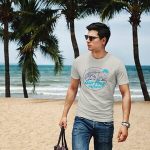 K&ouml;nnte beinhalten: Ein Mann tr&auml;gt ein hellgraues T-Shirt mit einer blauen und wei&szlig;en Grafik einer Welle und dem Text "Surfing Paradise". Er tr&auml;gt eine blaue Jeans und eine Sonnenbrille und geht an einem Sandstrand mit Palmen im Hintergrund entlang.