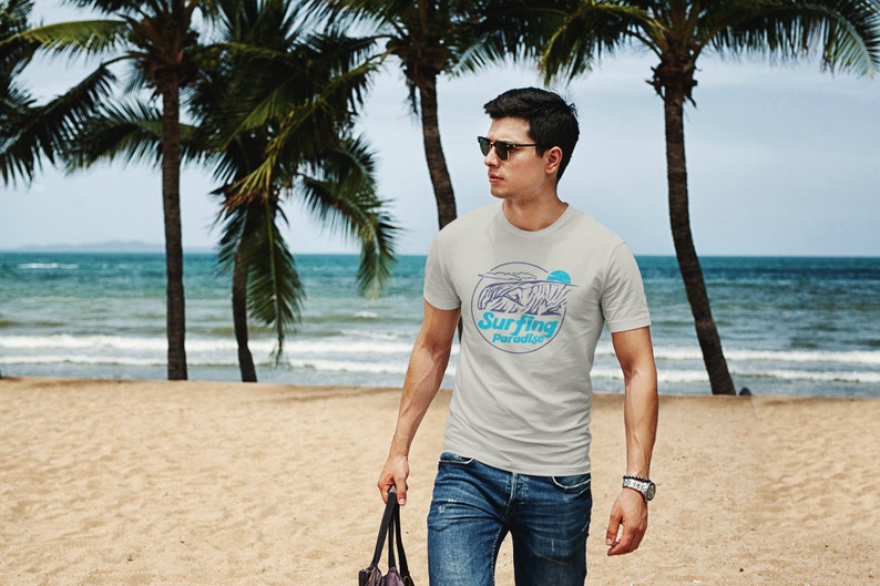 K&ouml;nnte beinhalten: Ein Mann tr&auml;gt ein hellgraues T-Shirt mit einer blauen und wei&szlig;en Grafik einer Welle und dem Text "Surfing Paradise". Er tr&auml;gt eine blaue Jeans und eine Sonnenbrille und geht an einem Sandstrand mit Palmen im Hintergrund entlang.