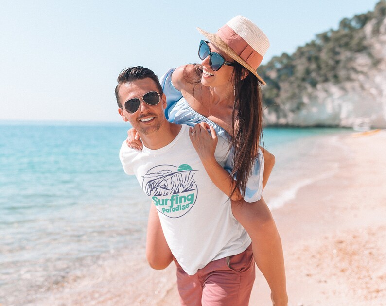 K&ouml;nnte beinhalten: Ein Paar am Strand, der Mann tr&auml;gt ein wei&szlig;es T-Shirt mit einer Grafik einer Welle und dem Text "Surfing Paradise". Die Frau tr&auml;gt ein blau-wei&szlig;es Kleid und einen Strohhut.