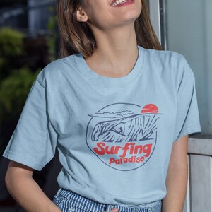 K&ouml;nnte beinhalten: Ein hellblaues T-Shirt mit einem Grafikdruck eines Surfers, der auf einer Welle reitet, mit einer roten Sonne und dem Text "Surfing Paradise".
