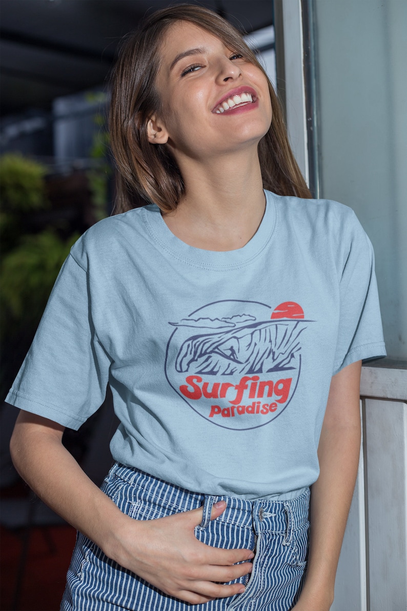 K&ouml;nnte beinhalten: Ein hellblaues T-Shirt mit einem Grafikdruck eines Surfers, der auf einer Welle reitet, mit einer roten Sonne und dem Text "Surfing Paradise".