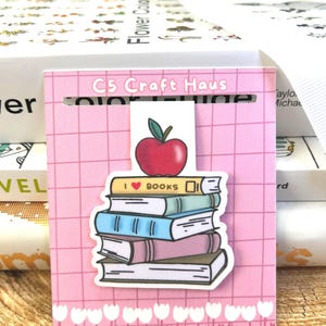 May include: A pink bookmark with a stack of books and a red apple on top. The bookmark has the text "I ❤️ BOOKS" on one of the books. The top of the bookmark has the text "C5 CRAFT HAUS".