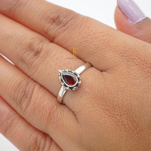 Peut inclure: Une bague en argent avec une pierre rouge en forme de larme sertie dans une bélière perlée. La bague est portée sur un doigt.