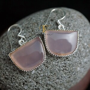 May include: A pair of silver earrings with a light pink, semi-circular gemstone. The earrings have a scalloped edge and a simple hook closure.