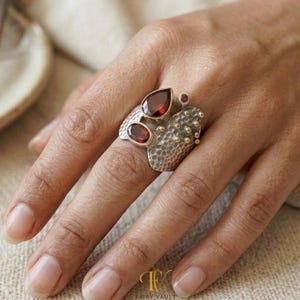 May include: A silver ring featuring two pear-shaped dark red gemstones and a textured surface with small gold-toned accents. The metal band has a hammered finish, and the design includes a small round red stone set near the larger pear-shaped gems.