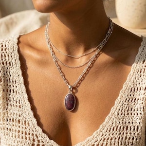 Peut inclure: Un collier en argent à trois rangs. Le rang inférieur comporte un grand pendentif ovale avec une pierre rouge foncé. Les deux autres rangs sont des chaînes délicates. La personne porte un haut en crochet crème.