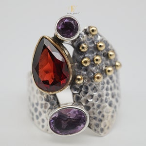 May include: A silver ring with a textured design features a red teardrop-shaped gemstone and two purple oval gemstones. The ring has gold accents.