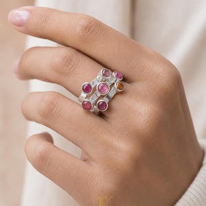 Può includere: Un anello in argento con tre fasce parallele collegate da una struttura metallica testurizzata. L'anello è incastonato con sei pietre preziose rotonde a taglio cabochon nei toni del rosa, magenta e arancione, disposte in modo sfalsato.