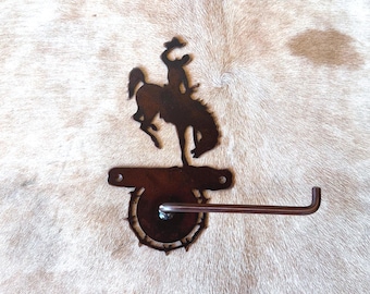 May include: A rustic metal toilet paper holder featuring a silhouette of a cowboy riding a bucking bronco. The holder is made of dark brown metal and has a horizontal bar for the toilet paper roll. The design is mounted on a textured background.
