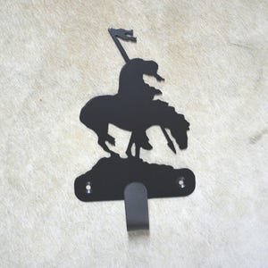 May include: Black metal wall hook shaped like a silhouette of a Native American riding a horse. The hook is attached to a wall with two screws.