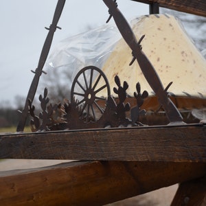 May include: A rustic wooden lampshade holder with a metal design featuring a wagon wheel and cacti. The holder is made of reclaimed wood and has a distressed finish. The lampshades are covered in a beige fabric with a speckled pattern.