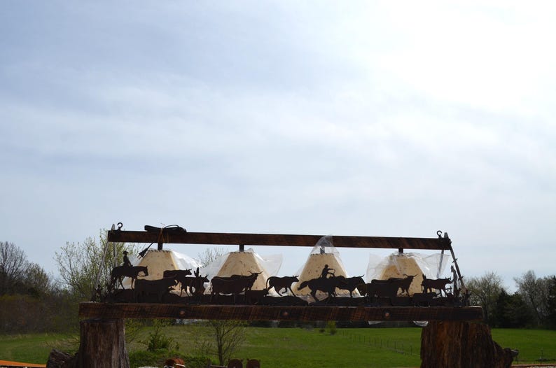 May include: A rustic metal chandelier with five light fixtures, featuring a silhouette of a cowboy riding a horse and cattle. The chandelier is mounted on a wooden beam.